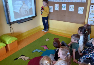 Chłopiec stoi z maskotką Krasnala Hałabały pod tablicą interaktywną na której wyświetlone jest zdjęcie Ratusza.